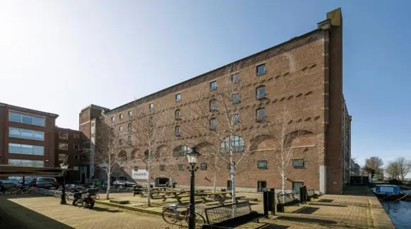 Rode bakstenen pakhuis aan de Hoogte Kadijk met kleine ramen en een plein met fietsen en bankjes op de voorgrond.